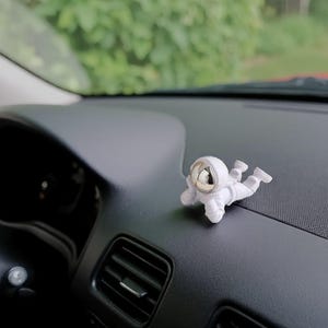 Op de afbeelding: Een kleine, witte astronautenfiguur met een reflecterend zilveren helm op een zwart dashboard in een auto. De astronaut ligt ontspannen, een decoratief item voor het interieur van een voertuig.