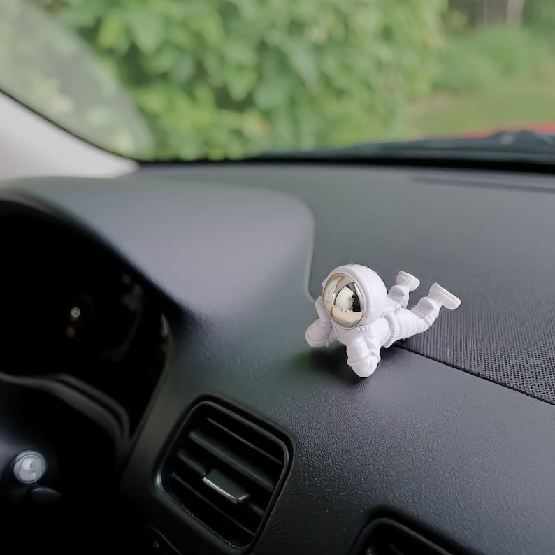 Op de afbeelding: Een kleine, witte astronautenfiguur met een reflecterend zilveren helm op een zwart dashboard in een auto. De astronaut ligt ontspannen, een decoratief item voor het interieur van een voertuig.