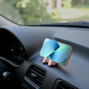 Op de afbeelding: Een smartphone met een goudkleurig frame wordt vastgehouden in een houten telefoonhouder. De telefoon toont een blauw en groen scherm met de tijd 09:41. De houder staat op een autodashboard.