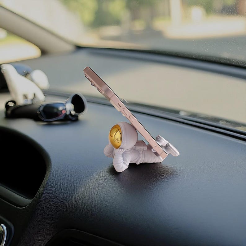 Op de afbeelding: Witte astronautvormige telefoonhouder met een goudkleurige helm, die een ros&eacute;gouden smartphone vasthoudt. De telefoonhouder staat op een autodashboard. Andere items op het dashboard zijn een zonnebril en een wit beeldje.