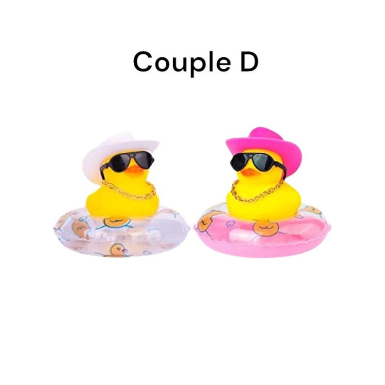 Op de afbeelding: Twee gele badeenden met zonnebrillen, gouden kettingen en cowboyhoeden, &eacute;&eacute;n witte en &eacute;&eacute;n roze. Ze drijven in opblaasbare ringen, &eacute;&eacute;n doorzichtig en &eacute;&eacute;n roze. De tekst "Couple D" staat erboven.