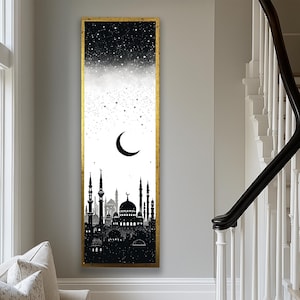 May include: A vertical framed art piece featuring a black and white silhouette of a cityscape with minarets and domes, under a crescent moon and starry sky. The artwork is encased in a gold frame, creating a striking contrast against the neutral wall.