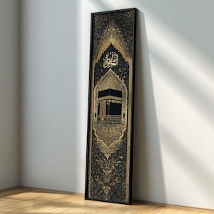 Op de afbeelding: Een ingelijst kunstwerk met een goud en zwart ontwerp, Arabische kalligrafie en een illustratie van de Kaaba. Het kunstwerk heeft een zwart frame en leunt tegen een witte muur en een houten vloer.