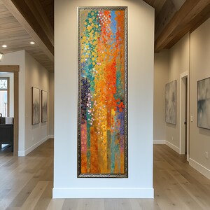 May include: A vertical abstract painting with a silver frame. The artwork features a vibrant array of colors, including orange, yellow, blue, and green, with a textured appearance. The painting is displayed in a hallway with light wood flooring.