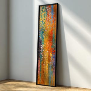 Può includere: Un'opera d'arte incorniciata, lunga e verticale, con un design astratto. L'opera presenta un vivace mix di colori, tra cui arancione, giallo, blu e verde, con un motivo strutturato e puntinato. La cornice è nera e l'opera è appoggiata a una parete bianca.