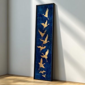以下が含まれることがあります： 深い青色の背景に、金色の鶴のシルエットが描かれた、額装された縦長の芸術作品。 黒いフレームに入っており、白い壁と木の床に立てかけられています。 鶴が飛んでいる様子を描いています。