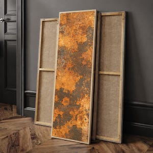 May include: A tall, rectangular canvas print featuring a textured, weathered surface with mottled shades of deep orange, rust, and dark charcoal gray. The artwork is mounted on a wooden frame, displayed alongside two empty, unprinted wooden frames.