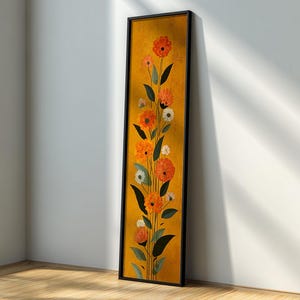Pode incluir: Uma obra de arte emoldurada com orientação vertical, apresentando flores. A imagem mostra flores laranja e brancas com folhas verdes sobre um fundo amarelo. A moldura é preta. A obra está encostada a uma parede branca.