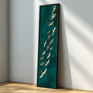 Puede incluir: Impresión artística enmarcada verticalmente que representa una bandada de grullas en vuelo. Las aves tienen alas blancas y doradas sobre un fondo verde azulado. El marco negro añade un toque moderno a la obra de arte, adecuada para la decoración del hogar.