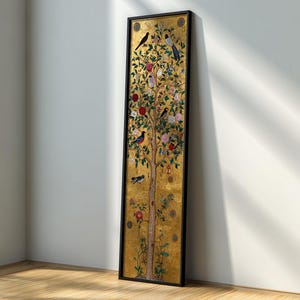 May include: A framed artwork with a golden background and a detailed tree design. The tree is decorated with colourful birds, red fruits, and pink flowers. The black frame contrasts with the gold, leaning against a white wall and wooden floor.