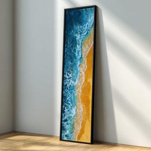 以下が含まれることがあります： 海岸の風景を描いた、額装された縦長の美術印刷物。鮮やかな青い海が、黄金色の砂浜と出会う様子が描かれています。黒いフレームが、ビーチをテーマにしたアートワークにモダンな雰囲気を加えています。
