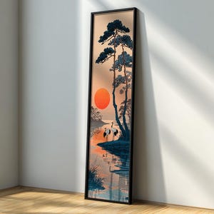 Puede incluir: Una impresión artística enmarcada que presenta un paisaje sereno con un sol naranja vibrante que se pone sobre el agua. Árboles silueteados y dos grullas se alzan en la orilla. La obra de arte tiene un formato vertical con un marco negro.