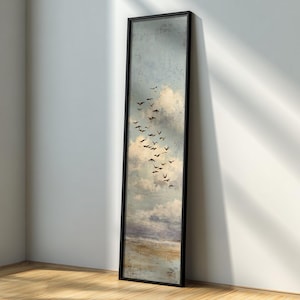 May include: A long, rectangular framed artwork featuring a cloudy sky with a flock of birds in flight. The artwork has a black frame and a muted color palette of blues, whites, and browns. The artwork is leaning against a white wall.