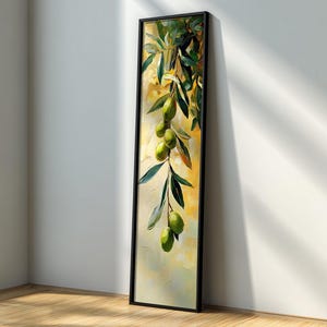May include: Vertical framed art print featuring an olive branch with green olives and leaves. The painting has a yellow and green background with a black frame. The artwork is leaning against a white wall and wooden floor.