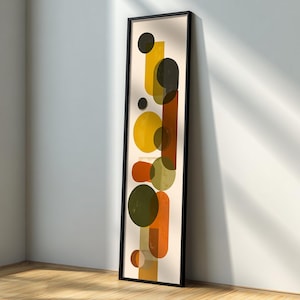 Puede incluir: Una impresión artística abstracta enmarcada con una composición vertical de formas geométricas. La obra de arte incluye círculos y rectángulos en tonos amarillos, naranjas y verde oliva sobre un fondo crema. El marco es negro.