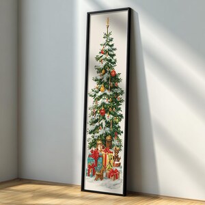 May include: A framed vertical print of a Christmas tree, decorated with ornaments and topped with a candle. Presents are at the base of the tree. The print is in a black frame and leans against a white wall.