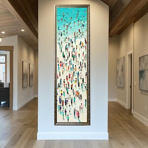 May include: Vertical framed artwork depicting a beach scene with numerous figures. The top shows a turquoise ocean with swimmers, transitioning to a sandy shore filled with people in various colors. The frame is a dark metallic color.