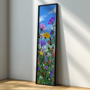 May include: A vertical framed painting of colorful wildflowers against a blue sky with fluffy white clouds. The flowers are in shades of pink, yellow, purple, and red, set against a green background. The frame is black.