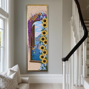 Può includere: Un dipinto rettangolare verticale in cornice dorata che raffigura una finestra aperta su un mare blu e scogliere. La finestra è incorniciata da glicini viola, girasoli gialli e iris azzurri su una parete beige dalla superficie ruvida.