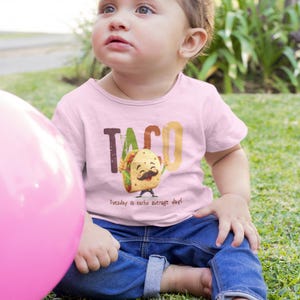 Peut inclure: Un t-shirt rose pour enfant avec un dessin animé de taco et le texte "Taco Tuesday is nacho average day!"