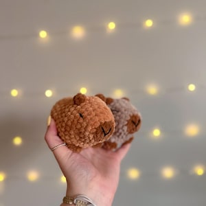 May include: Two crocheted capybara plushies. One is a warm brown color, the other is a light grey. Both have small, dark brown eyes and noses. The plushies are held up against a background of soft, out-of-focus lights.