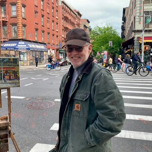 May include: A man wearing a green jacket and a black baseball cap stands in front of a wooden easel with two paintings on it. The paintings are of a street scene with buildings and a crosswalk. The man is smiling and looking at the camera. The background is a city street with buildings and trees.