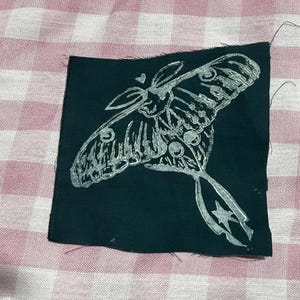 May include: A rectangular black fabric patch featuring a screen-printed silver ink illustration of a moth. The moth design includes detailed wing patterns, a small heart shape above its head, and a star symbol on its long, trailing tail.