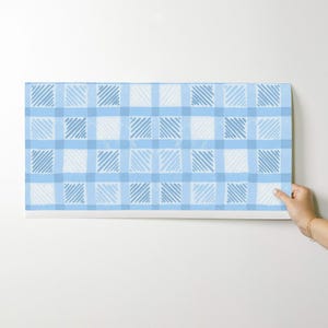 May include: A rectangular piece of light blue and white plaid fabric. The plaid pattern features a grid of light blue lines with alternating squares of solid and diagonally striped light blue. The fabric is being held up against a white background.