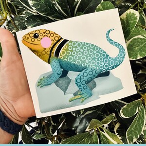 May include: A colorful illustration of a lizard with a yellow, green, and blue patterned body. The lizard is standing on a white rock with a shadow underneath.