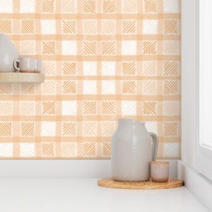 May include: A kitchen scene featuring a light orange and white plaid wallpaper. A white countertop holds a wooden tray with a gray pitcher and a small pink cup. A wooden shelf displays a white pitcher and two small cups.