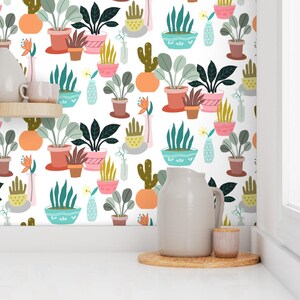 May include: White wallpaper with a repeating pattern of potted plants in various colors. The plants include cacti, succulents, and leafy plants in terracotta, pink, and blue pots. A white pitcher, small cup, and wooden tray are in the foreground.
