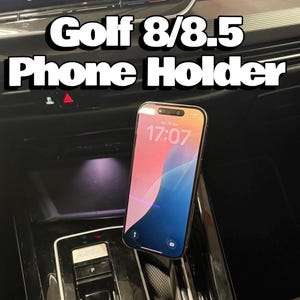 Peut inclure: Support de téléphone pour modèles Golf 8/8.5. L'image montre un smartphone affichant l'heure, maintenu dans la console d'une voiture. Le texte "Golf 8/8.5 Phone Holder" est affiché au-dessus du téléphone.