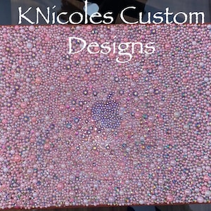May include: A pink and pearl-covered laptop cover with the Apple logo in the center. The cover is adorned with various sizes of pink pearls and iridescent rhinestones. The text "KNicoles Custom Designs" is at the top.