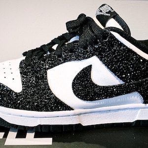 Può includere: Sneakers bianche e nere con un design scintillante e glitterato. Le scarpe presentano una base bianca con dettagli neri, inclusi i lacci e il logo. Le aree della punta e del tallone sono ricoperte di glitter neri.