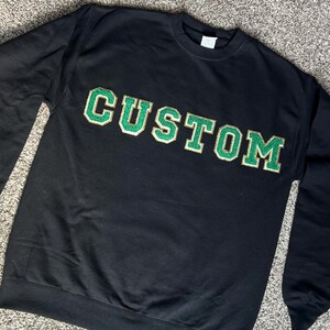 Könnte beinhalten: Schwarzer Pullover mit dem Wort "CUSTOM" in grünen und goldenen Buchstaben.