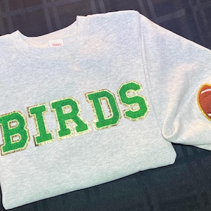 以下が含まれることがあります： グレーのスウェットシャツに、緑と金のグリッターで「BIRDS」の文字が書かれています。右袖には、白い紐のついた茶色のフットボールがあります。