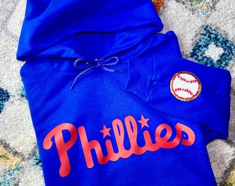 Może przedstawiać: Royalnie niebieski sweter z kapturem z czerwonym logo "Phillies" i białą piłką baseballową z czerwonymi szwami na rękawie.