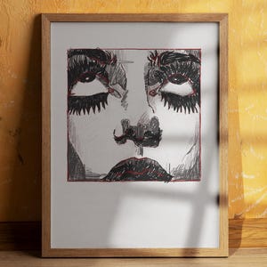 May include: A framed art print depicting a stylized face. The drawing is mainly black and white, with red detailing on the facial features. The eyes have thick lashes, and the lips are full. The frame is made of light-coloured wood.