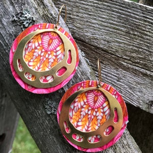 Pode incluir: Um par de brincos de argola dourados com um design floral de tecido rosa e laranja. O tecido é em camadas e a argola dourada tem um design recortado.