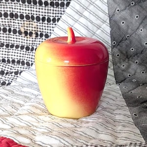 May include: A ceramic apple-shaped container with a lid. The container is yellow at the base, transitioning to red at the top, with a small stem. The container is sitting on a patterned quilt.