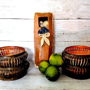 May include: A tall, rectangular brown ceramic wall pocket featuring a small skull figure in an upper cutout, decorated with a blue flower and a white bow. Two dark, slatted metal candle holders with warm interior light flank three green limes.
