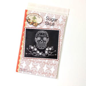 May include: A package featuring a Sugar Skull design. The design is a detailed white embroidered skull on a black background, surrounded by floral elements. The package has a red and orange border and is sealed in clear plastic.
