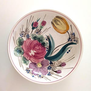 May include: A round, off-white ceramic bowl with a hand-painted floral design. The bowl features a large pink rose, a yellow tulip, and other flowers in shades of purple, green, and blue. A thin pink line borders the rim.