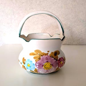May include: A white ceramic basket with a handle, adorned with hand-painted floral designs in shades of blue, purple, yellow, and brown. The rim and handle are outlined in green. This decorative basket is suitable for home decor.