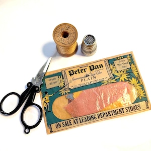 May include: A vintage sewing kit with a wooden spool of thread, a metal thimble, black-handled scissors, and a Peter Pan fabric label. The label reads "Guaranteed Fast Color Plain".