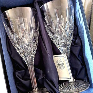 May include: Two crystal wine glasses with intricate diamond-cut patterns, presented in a blue box with navy satin lining. The glasses have long stems and wide bowls. A tag with the "STUART CRYSTAL" logo is visible.