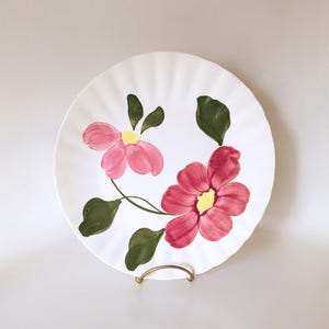 May include: White ceramic plate with a scalloped edge, featuring hand-painted pink flowers with yellow centers and green leaves. The plate is displayed on a gold-colored metal stand.