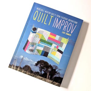 May include: A book titled "Quilt Improv" by Lucie Summers. The cover features a colorful quilt design hanging on a clothesline against a blue sky background. The book's title is in large, stylized letters.