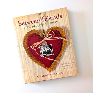 May include: A book titled "between friends craft projects to share" with a red heart-shaped decoration. The heart has a photo and button. The book cover has a wood-like texture and the author's name is Charlotte Lyons.