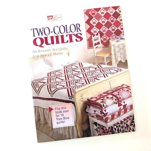 May include: A book titled "TWO-COLOR QUILTS" featuring ten romantic red quilts by Nancy J. Martin. The cover displays a red and white quilt design. A stack of quilts and a framed quilt are also visible.